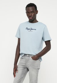 Gaiši zils kokvilnas T-krekls ar "Pepe Jeans London" logo tumši zilā un sarkanā krāsā. Apvienots ar gaiši pelēkiem džinsiem, demonstrēts uz modeļa.