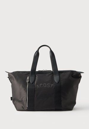 Bolso tote negro de Lacoste con dos asas, cremallera superior, un bolsillo vertical con cremallera en la parte delantera y un logotipo discreto.