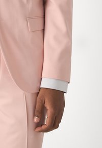 Completo rosa chiaro con rever a punta e una tasca, con un polsino di camicia bianca che spunta dalla manica, tessuto testurizzato.