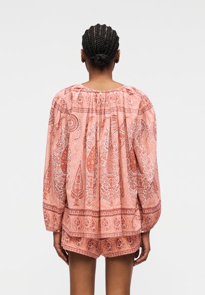 Femme avec un chignon tressé portant un chemisier ample à manches longues rose à motif cachemire et un short assorti, debout de dos sur un fond uni.