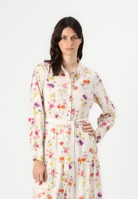 Rochie florală într-o nuanță deschisă de bej, cu un decolteu cu nasturi, mâneci lungi, talie prinsă cu un cordon și o fustă în straturi cu modele colorate de flori.
