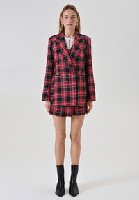 Blazer-Kleid in rot-schwarz kariert mit doppelter Knopfleiste, Reverskragen und plisseeartigem Rock. Kombiniert mit schwarzen Knöchelstiefeln.