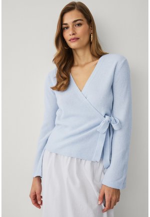 Kobieta o długich brązowych włosach, ubrana w jasnoniebieski sweter zapinany w pasie oraz białą spódnicę, stojąca na jednolitym tle.