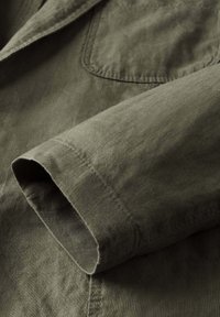 Nahaufnahme eines olivgrünen Langarmshirts, das die Manschette und eine Brusttasche mit sichtbaren Nähten zeigt.