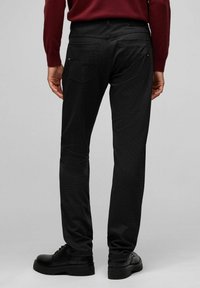 Jeans noirs avec une finition texturée, design à cinq poches et coupe ajustée. Portés avec des bottes noires à lacets, mettant en valeur les détails de la poche arrière.