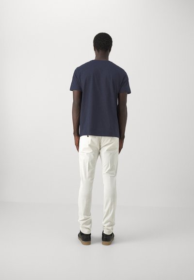 T-shirt di cotone navy con maniche corte, abbinata a pantaloni bianchi slim-fit e sneakers nere con suola in gomma. È in piedi di spalle.