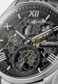 Zegarek mechaniczny z stalową kopertą, czarną szkieletową tarczą, srebrnymi znacznikami godzin i złotymi akcentami. Posiada skomplikowany design zębatek.