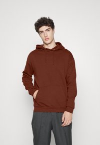 Brown fleece hoodie with a kangaroo pocket, drawstring hood, relaxed fit, and ribbed cuffs, paired with gray trousers.