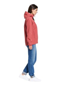 Lichtroze waterdichte jas met een capuchon, elastische manchetten en twee zijzakken, gedragen met blauwe jeans en witte schoenen.