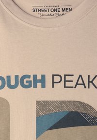 Ljusbeige t-shirt med blå och mörkgrå grafisk textöverlägg, med abstrakta geometriska former och texturerade mönster. Rund halsringning.