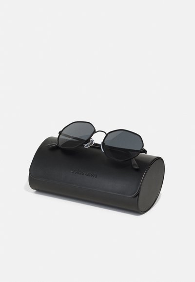 Black octagonal sunglasses with dark lenses resting on a cylindrical black leather case embossed with "Giorgio Armani."