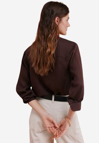 Bruine langarmshirt van een textiele stof, met een rugpand en opgerolde mouwen, gedragen met lichtbeige corduroy broeken.