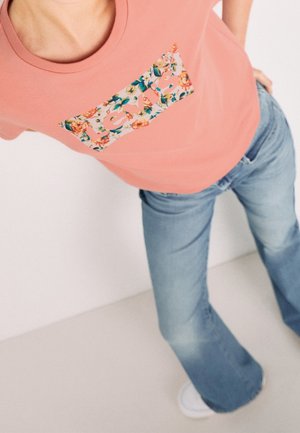 Personne portant une chemise rose fleurie "Levi's" et un jean bleu, debout les mains sur les hanches sur fond blanc.