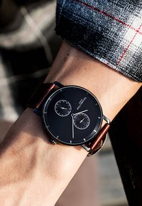 Montre analogique noire avec deux sous-cadrans, accents argentés et un bracelet en cuir marron. Cadran rond, design minimaliste et repères horaires clairs.