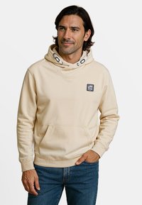 Sweatshirt à capuche couleur crème avec une poche frontale, des poignets côtelés et un patch logo sur la poitrine gauche. Tissu lisse et décontracté.