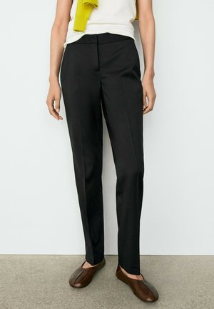 Personne portant un pantalon noir droit, un haut blanc avec un pull jaune drapé sur les épaules, et des chaussures plates marron, debout sur un sol gris.