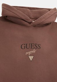 Felpa cappuccio marrone realizzata in tessuto morbido, con tasca a marsupio. Sul davanti è presente il logo "GUESS" in nero con un emblema triangolare sottostante.