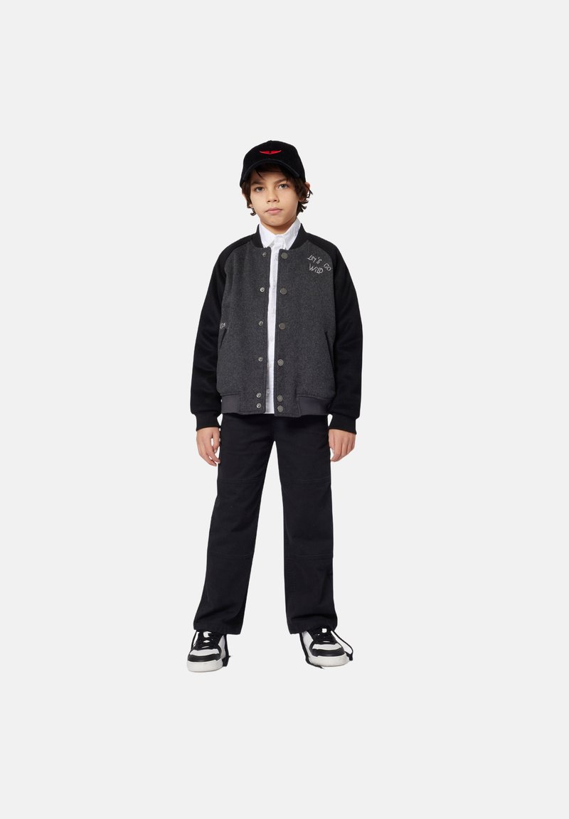 Zwart en grijs varsityjack met drukknopen, gecombineerd met een zwarte pet en zwarte broek; voorzien van geborduurde tekst en een gestructureerde afwerking.