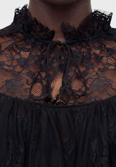 Άτομο με σκουρόχρωμο τόνο δέρματος που φοράει διαφανές μαύρο δαντελωτό μπλουζάκι με λουλουδένια σχέδια και δεμένη φιόγκο στον ψηλό γιακά.