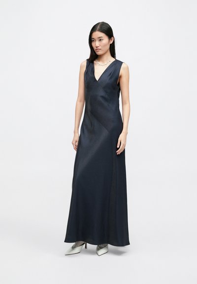 Μακριά φόρεμα από σατέν σε ναυτικό μπλε, με βαθύ V-λαιμό, χωρίς μανίκια και απλή υφή, συνδυασμένο με λευκά ψηλοτάκουνα παπούτσια.