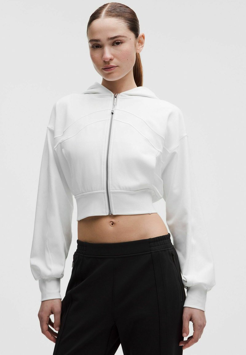 Donna con capelli castani lisci raccolti in una coda di cavallo, indossa una felpa bianca corta con cerniera e pantaloni neri a vita alta su uno sfondo semplice.