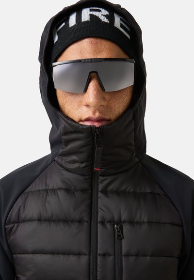 Sort isoleret dunjakke med høj krave, lynlåslommer og kontrasterende skulderpaneler. Mørk hue med teksten "FIRE" og spejlglas solbriller.
