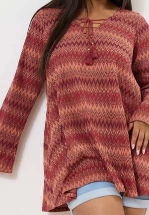 Femme portant une tunique à manches longues avec un motif zigzag rouge et rose et un col à nouer avec des pompons, associée à un short bleu clair retroussé.