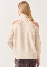 Pull en maille beige clair avec col haut, épaules dégagées et texture côtelée ; présente une coupe décontractée et des épaules tombantes.