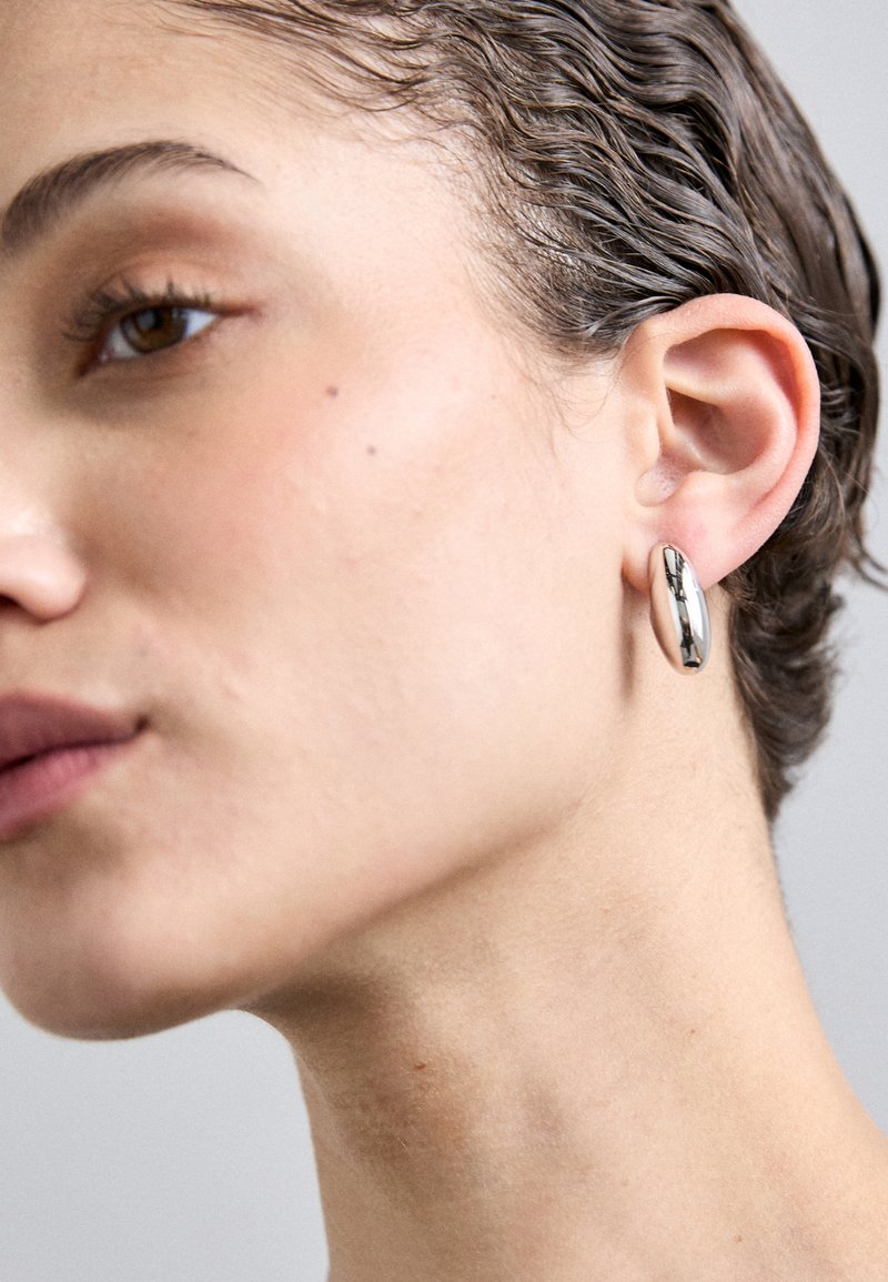 Srebrni metalni naušnici u obliku obruča s glatkom, reflektirajućom površinom, ovalnog oblika i suptilnom debljinom, prikazani u krupnom planu.