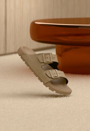 Beige Slidesandale mit zwei verstellbaren Schnallenriemen, dicke strukturierte Sohle, schwebt über einem beigen Teppich in der Nähe eines runden braunen Tischfußes.