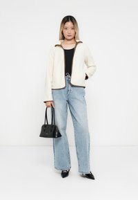 Creme-kleurige fleece jas met bruine accenten, gecombineerd met lichtblauwe high-waisted jeans. Model houdt een kleine zwarte handtas vast en staat tegen een witte achtergrond.