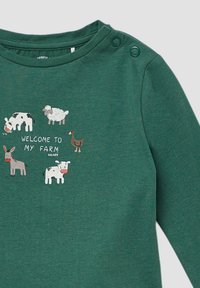 Grünes langärmliges Baumwollshirt mit Tierillustrationen: Kühe, Schafe, Esel und Ente, mit dem Text "WILLKOMMEN IN MEINER FARM". Zwei Druckknöpfe an den Schultern.
