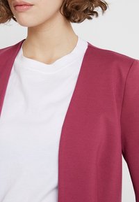 Bordeauxfärgad cardigan lager över en vit t-shirt, med ett mjukt, lättviktigt material och öppen framsidesdesign.