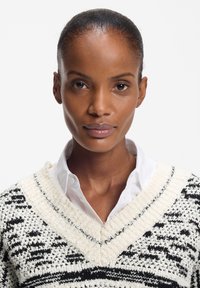 Γυναίκα με λείο μαζεμένο πίσω μαλλί, φοράει λευκό πουκάμισο με γιακά και πλεκτή μαύρη και λευκή μπλούζα με μοτίβο, κοιτάζοντας ευθεία μπροστά.