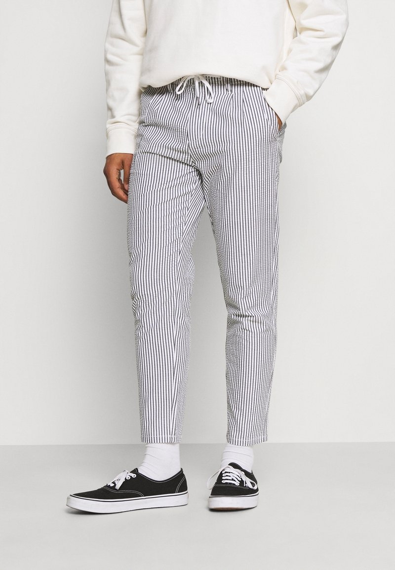 Stribede bukser i hvid og marineblå, lavet af et stof med en glat tekstur. Tapered fit med elastisk talje og sidelommer. Båret med sorte sko.