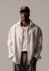 White oversized button-up shirt with a chest pocket, worn over a white ribbed tank top, paired with gray pants and a gray cap. Gold necklaces visible.