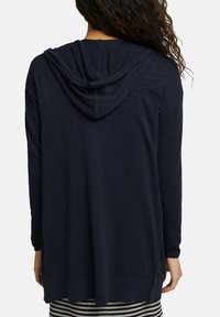 Marineblauwe hoody cardigan met lange mouwen, zachte textuur en een relaxte pasvorm; heeft een gebogen zoom en geribbelde manchetten. Gestreepte rok zichtbaar daaronder.