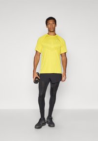 Camiseta atlética amarilla con patrón texturizado, pantalones cortos negros, mallas negras y zapatillas. Sosteniendo una botella de agua, de pie en una pose neutral.