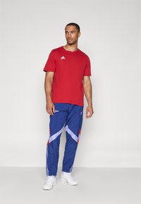 Červené bavlněné tričko s krátkým rukávem, doplněné modrými sportovními kalhotami s červenými a bílými detaily. Bílá obuv dotváří vzhled.