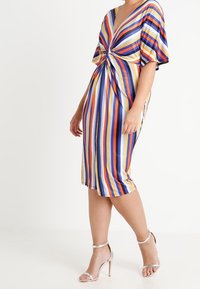 Robe midi rayée multicolore avec un profond décolleté en V, une taille cintrée et des manches courtes. Le tissu semble lisse et léger. Talons argentés.
