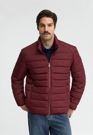 Uomo con baffi che indossa una giacca trapuntata color bordeaux sopra una camicia scura e jeans blu, in piedi davanti a uno sfondo chiaro uniforme.