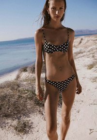 Femme portant un bikini à pois noirs et blancs debout sur une plage de sable près d'une mer calme sous un ciel clair.