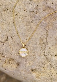 Guldhalsband med en cirkulär hängande pendant med "amor" utskuret, mot en pärlemorskimrande bakgrund, hängande på ett fint kedja.