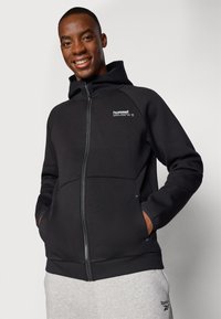 Uomo sorridente che indossa una felpa con zip nera con il logo "hummel" e pantaloni della tuta grigio chiaro con il logo Reebok, mani nelle tasche, su uno sfondo semplice.