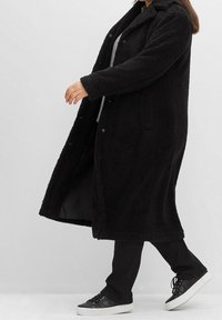 Musta, pitkä, pörröinen takki, joka on valmistettu teksturoidusta materiaalista. Takissa on nappikiinnitys, sivutaskut ja rento istuvuus. Takki on puettu harmaan yläosan ja mustien housujen päälle.