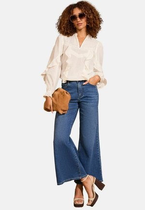 Blouse blanche à volants, jeans larges bleus, pochette en daim marron et talons compensés beiges. De grandes lunettes de soleil complètent la tenue.