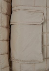 Beige quiltet pufferjakke med en rektangulær klaplomme på forsiden, med en glat tekstur og kontrasterende syninger.