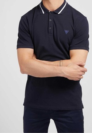 Homme portant un polo bleu marine à rayures blanches sur le col et un pantalon foncé, bras croisés, montrant un tatouage au poignet.