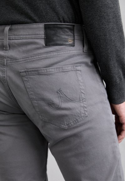 Pantalones de algodón grises que cuentan con un bolsillo trasero con un patrón de costura decorativa y una etiqueta de cuero negra con un logo circular.