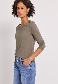 Kvinna med lockigt hår iförd en långärmad olivgrön skjorta och blå jeans, som står med en hand i fickan framför en enfärgad bakgrund.